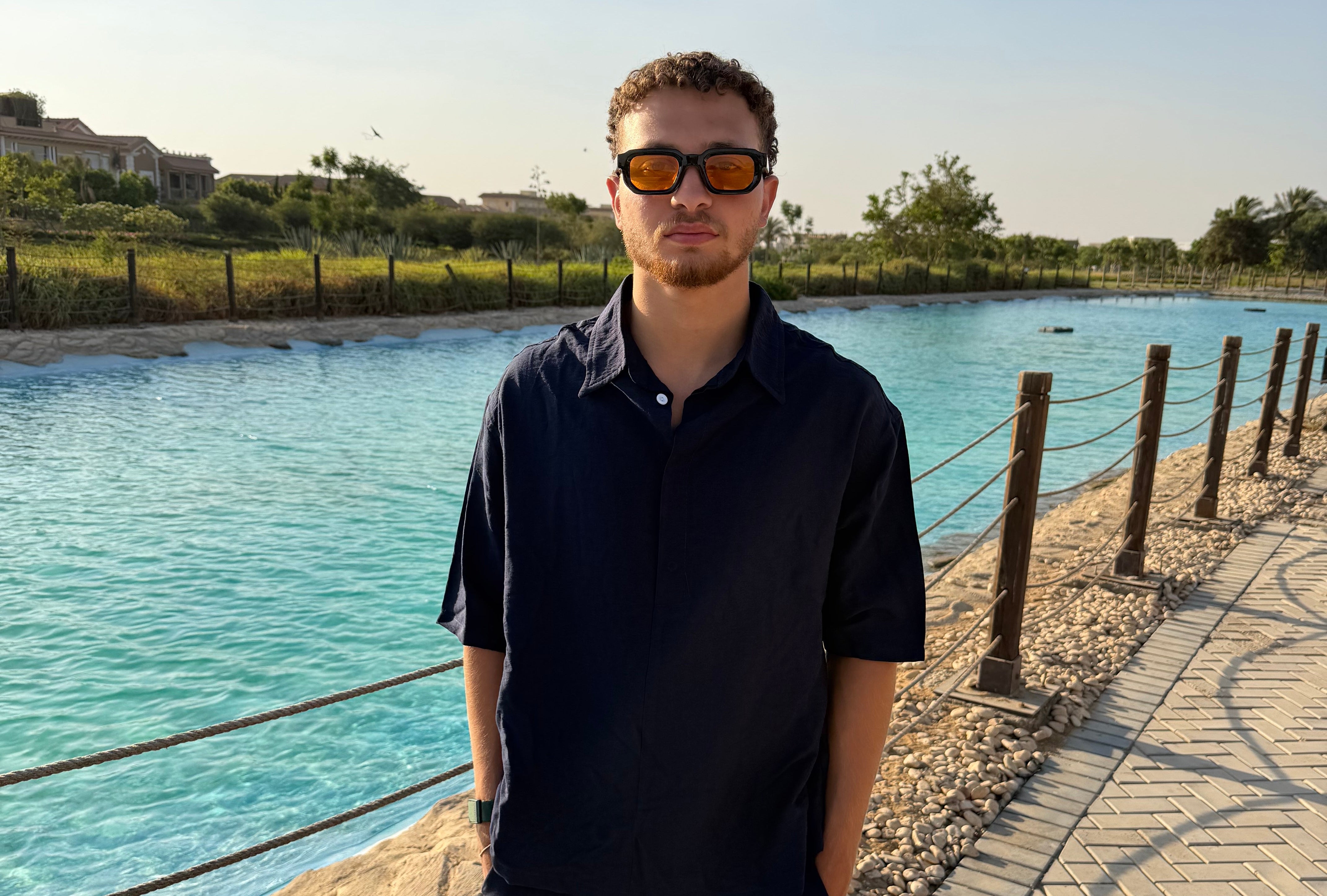 A man wearing a dark navy blue polo shirt and matching trousers stands outdoors on a paved walkway next to a body of water, wearing orange-tinted sunglasses.